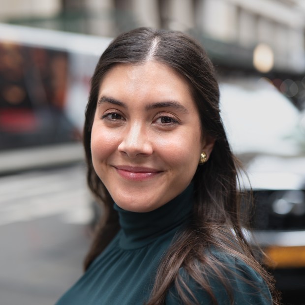 A woman with brown hair wearing a green turtleneck looks at the camera with a small smile on her face.